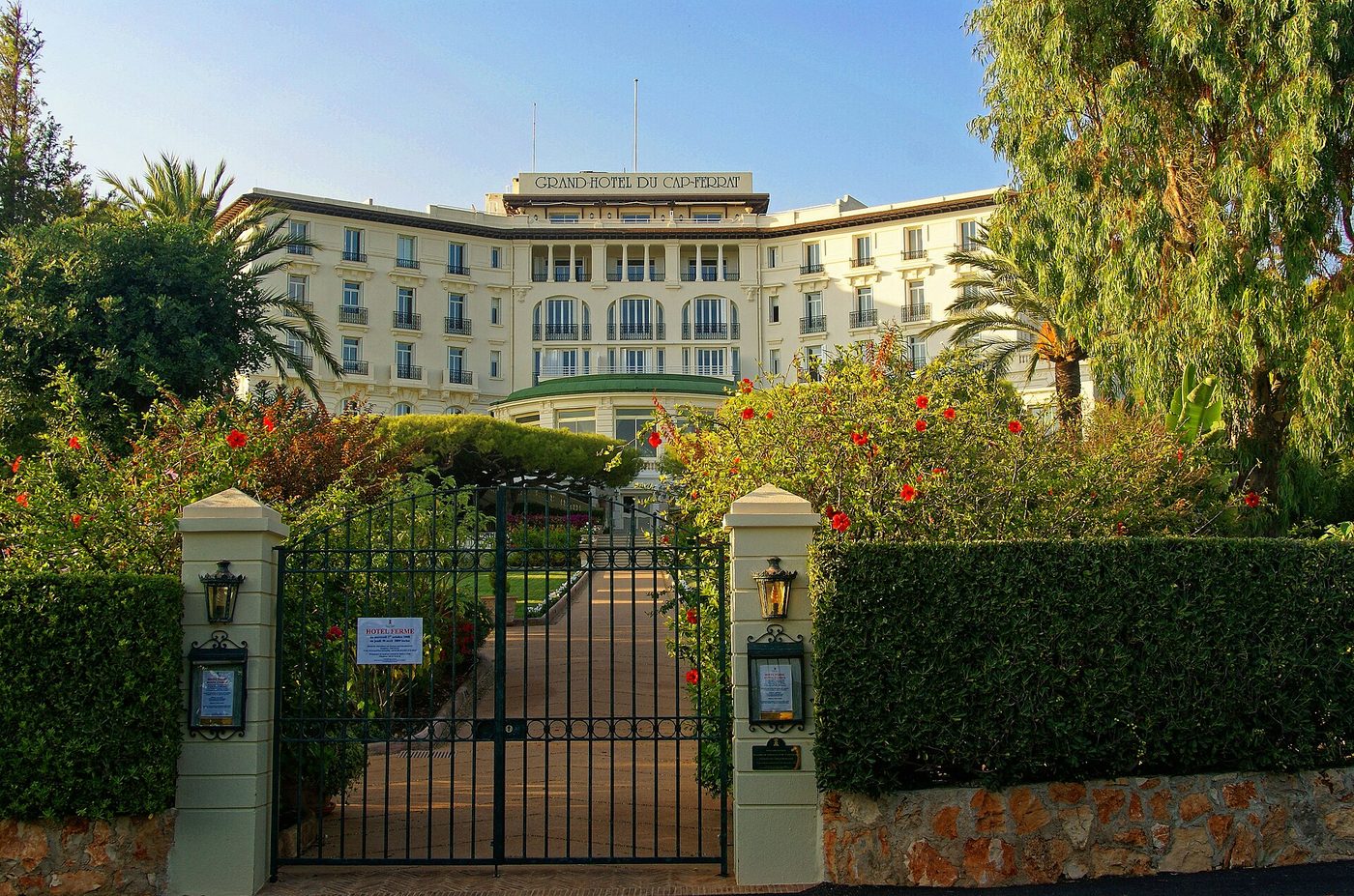 Le Grand-Hôtel du Cap-Ferrat vu depuis sa grille d'entrée, entre les palmiers et les massifs fleuris