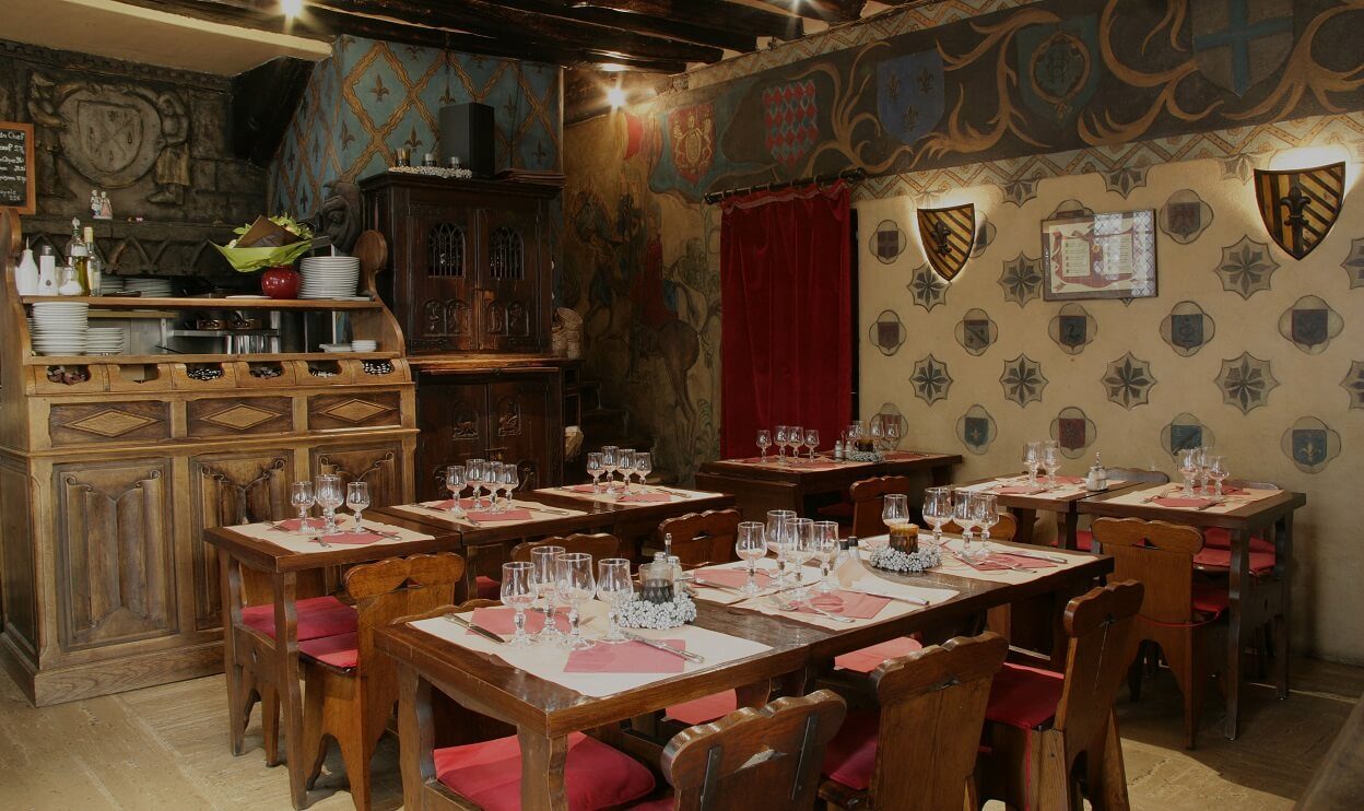La salle d'Aux Anysetiers du Roy, tables nappées de rouge, blasons au mur, tentures et comptoir de bois