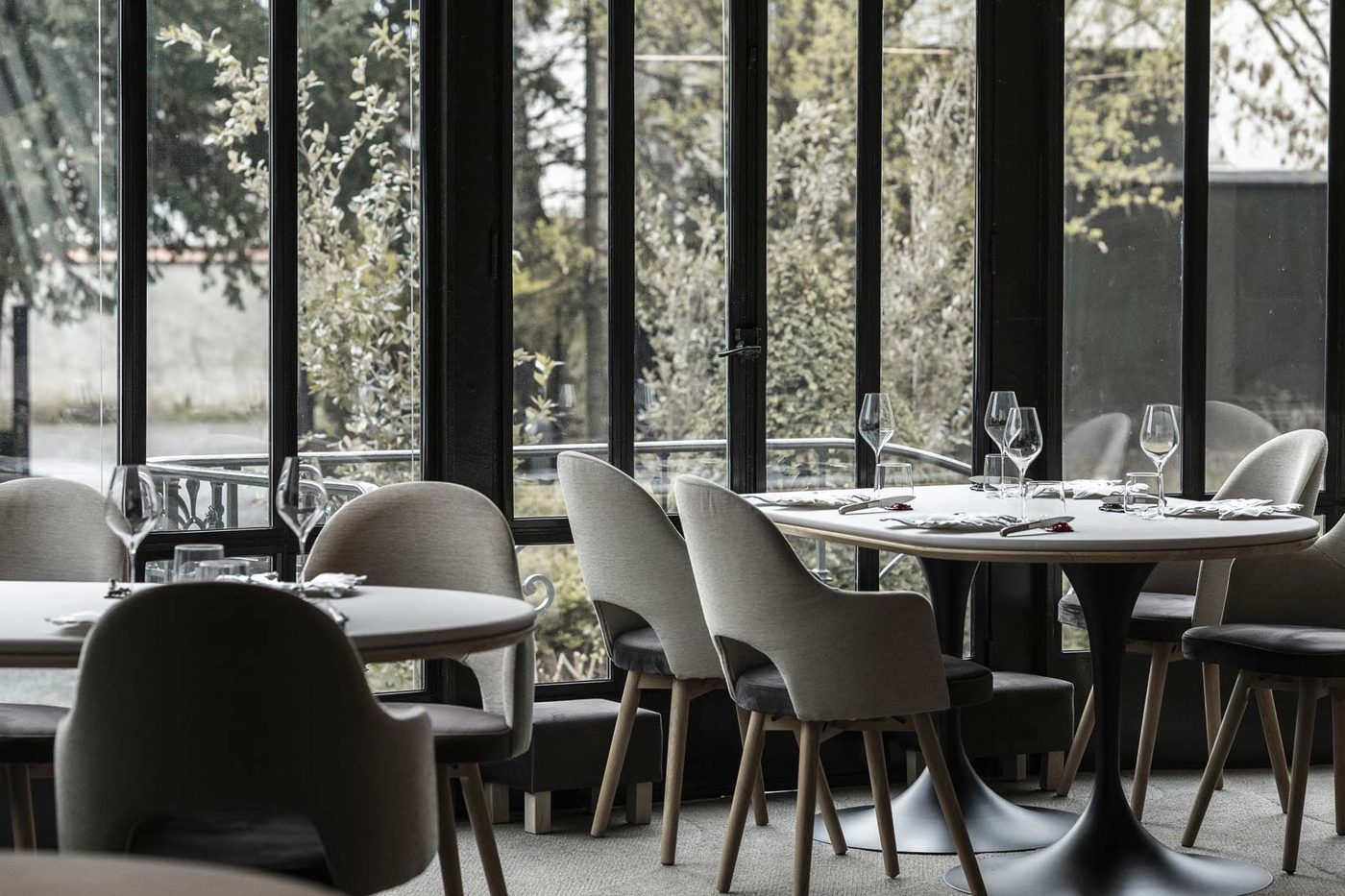 La salle d'Arbane : tables rondes nappées de blanc, fauteuils gris clair et grandes baies vitrées sur des arbres