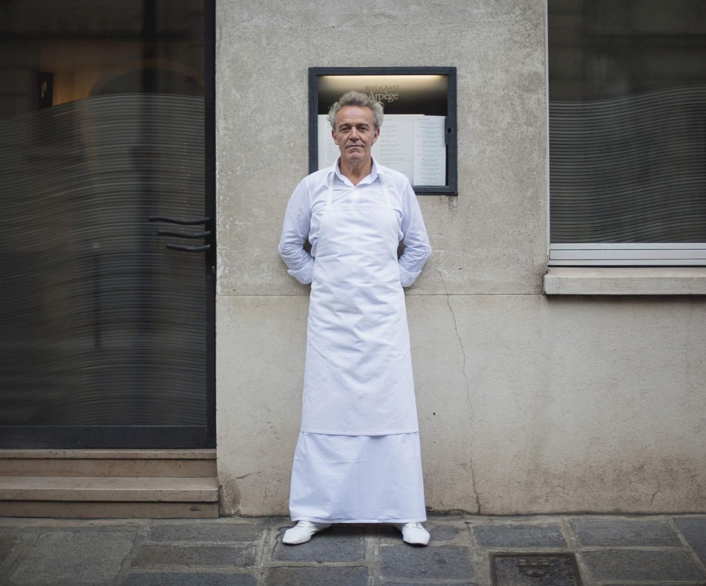 Alain Passard en tablier blanc devant la façade d'Arpège, rue de Varenne, sous la vitrine où est affichée la carte