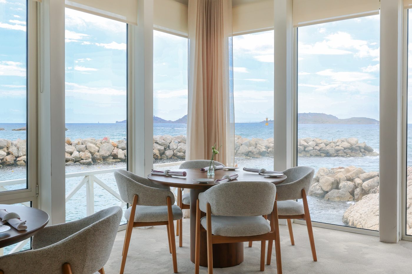 La salle d'Auffo, table ronde en bois foncé et fauteuils gris devant de grandes baies vitrées sur les rochers et la mer