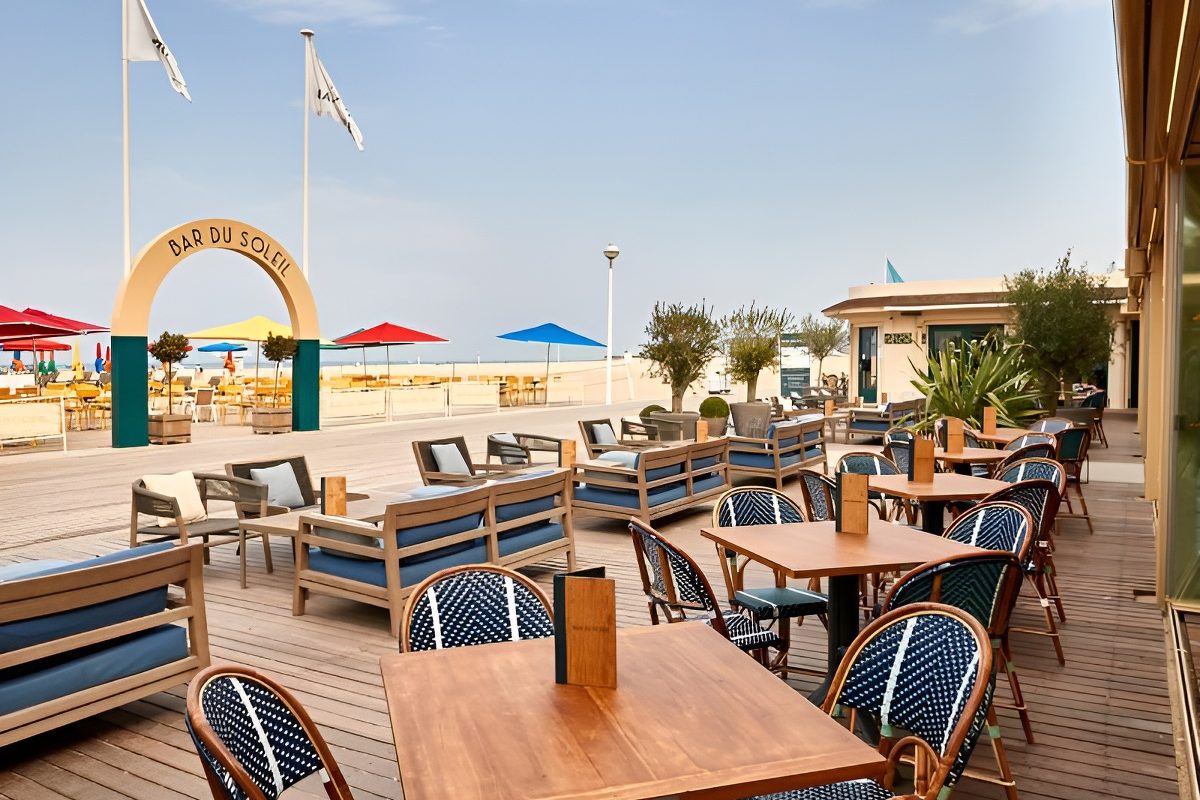 La terrasse en bois du Bar du Soleil, tables et fauteuils face à la plage, parasols colorés et arche à son nom