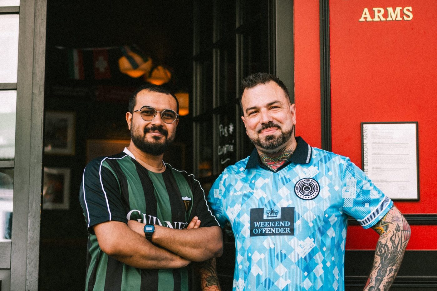 Deux hommes devant la devanture rouge du Blossom Arms, sous l'enseigne dorée du pub
