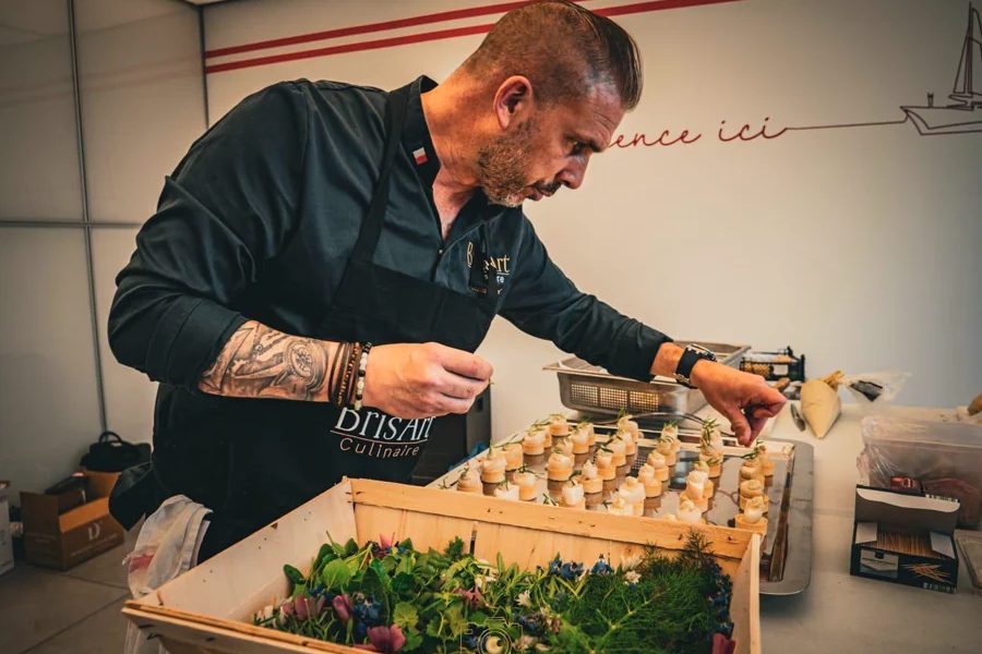 Guillaume Brisard penché sur un plateau de bouchées colorées, une caisse d'herbes fraîches devant lui