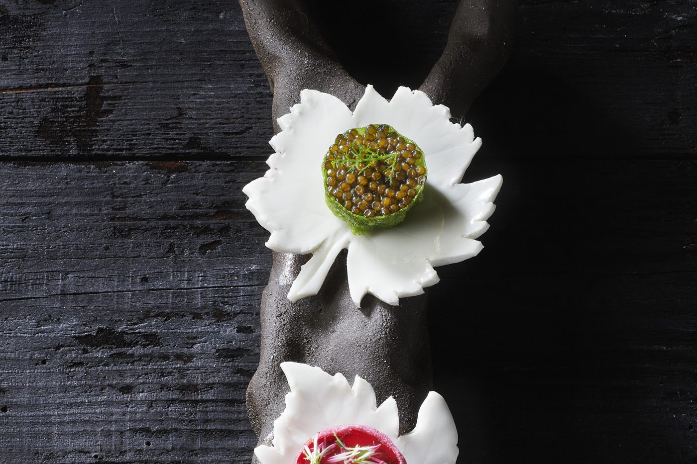 Une bouchée en forme de fleur blanche garnie de caviar, posée sur un bois sombre, au Chabichou