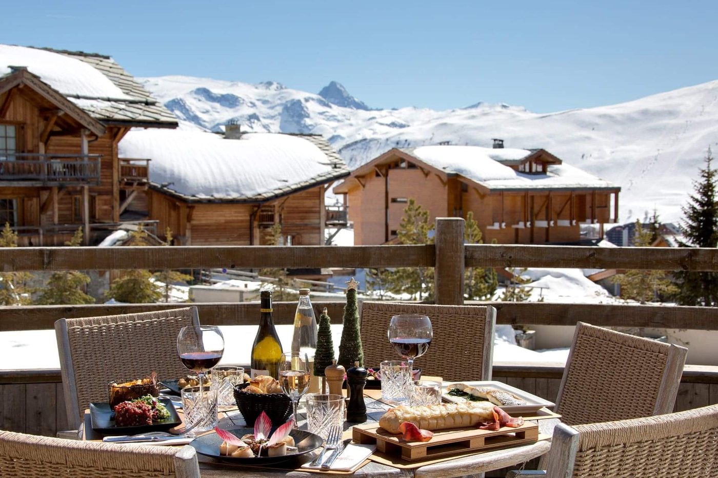 Une table dressée sur la terrasse enneigée du Chamois d'Or, planche de charcuterie et vin rouge, chalets et sommets blancs en arrière-plan