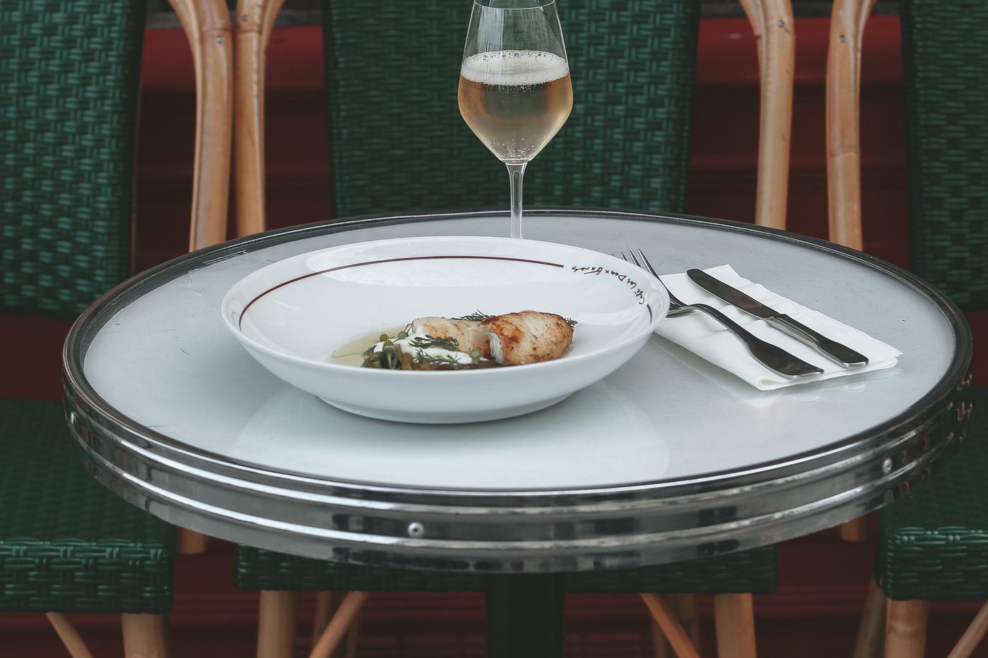 Un guéridon de bistrot cerclé de chrome en terrasse du Café Les Deux Gares, une assiette et un verre de vin