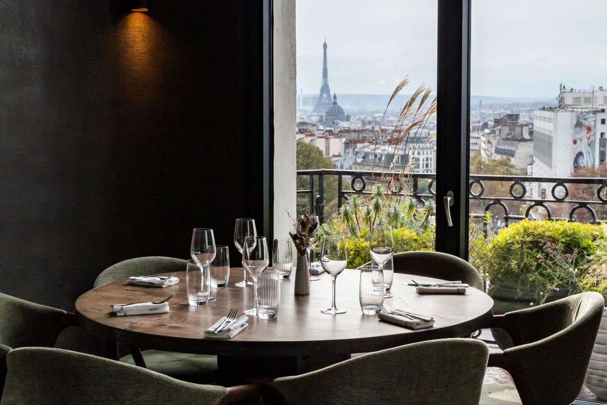 Une table ronde d'Edmond devant une grande baie à balcon de fer forgé, la tour Eiffel au loin sur les toits