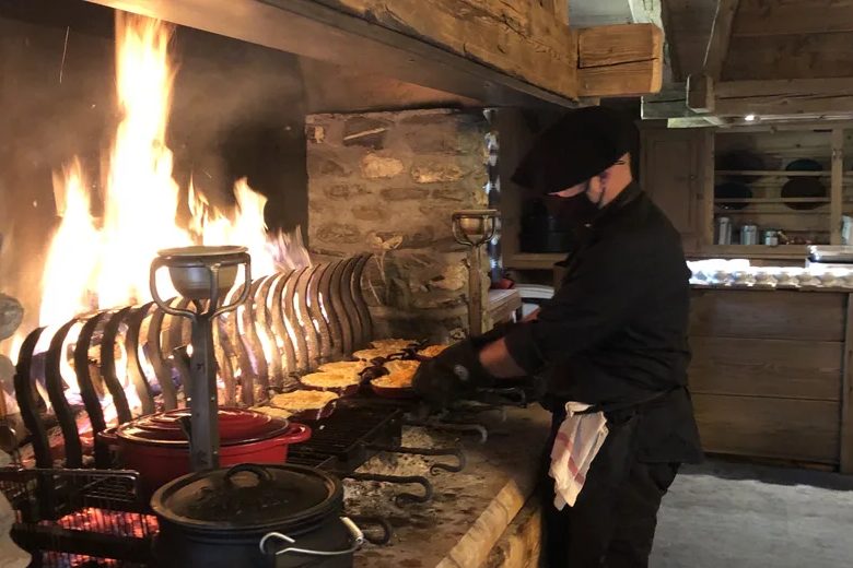 Un cuisinier devant la grande cheminée des Fermes de Marie, broches de viande rôtissant au feu