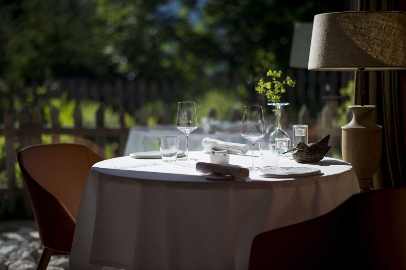 Une table ronde nappée de blanc dressée pour deux à Flocons de Sel, fauteuils couleur cognac et lampe en bois, devant le jardin
