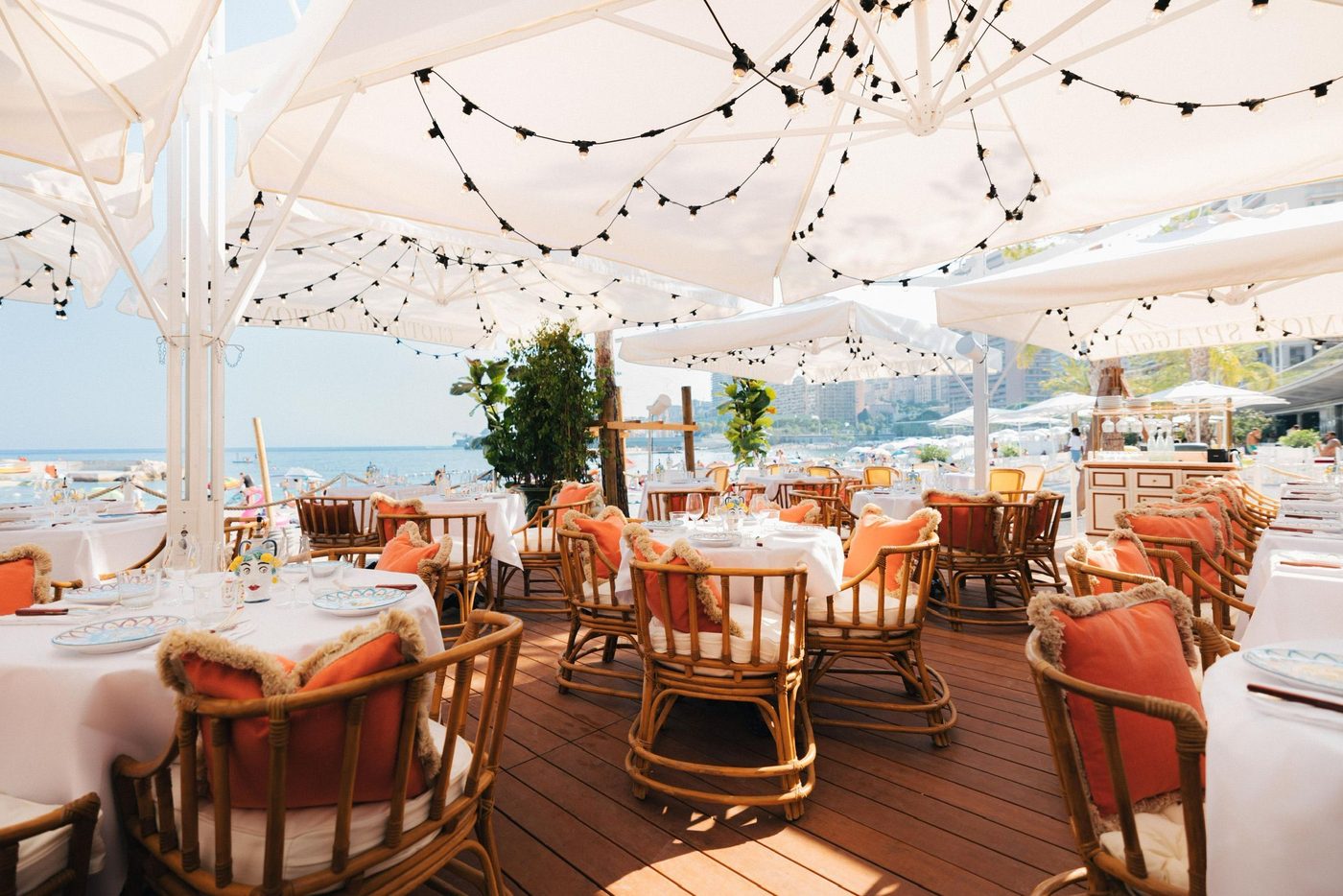 La terrasse de Giacomo sur la plage du Larvotto, fauteuils en rotin, coussins orange, parasols blancs et guirlandes
