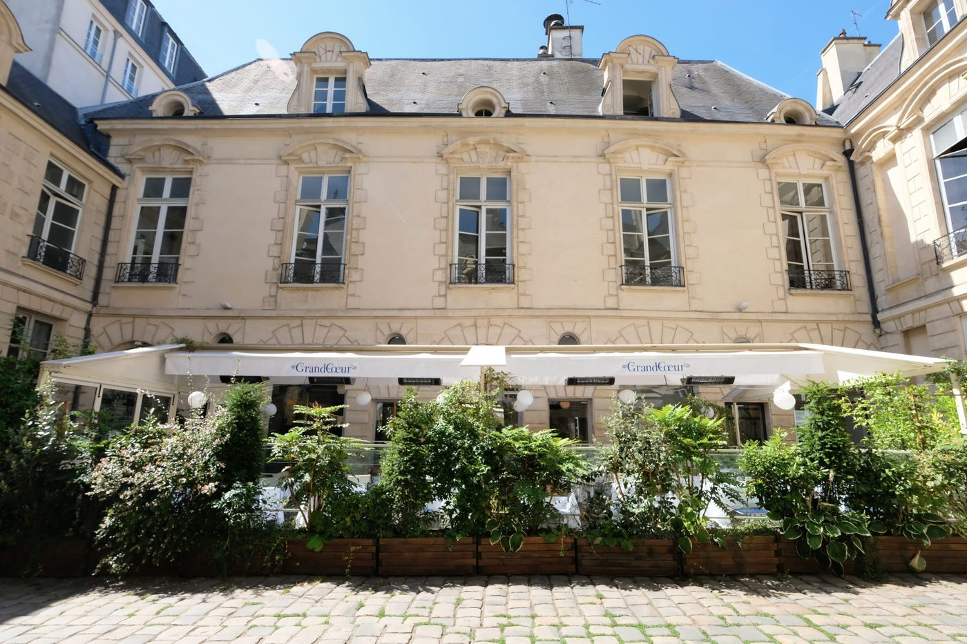 La terrasse de GrandCœur sous une toile blanche, dans la cour pavée bordée de façades du XVIIe siècle