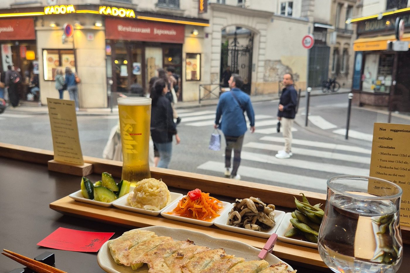 Une assiette de gyozas grillés, des petits plats et une bière sur un comptoir de bois face à la vitrine