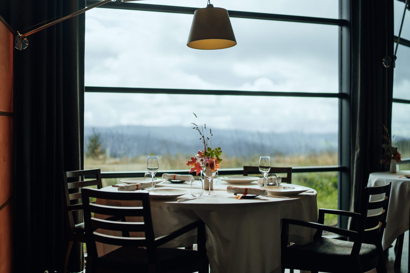 Une table ronde nappée de blanc au Restaurant Marcon, devant une grande baie vitrée ouverte sur la prairie et les montagnes