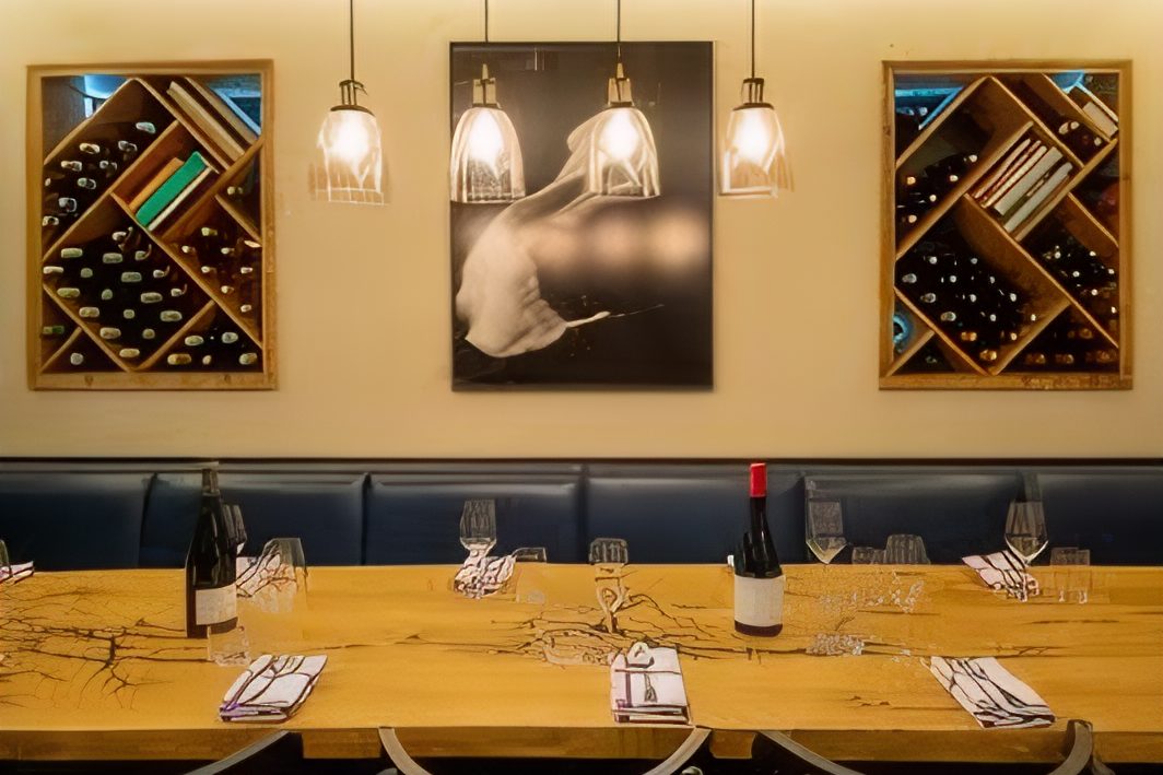 La salle du Café des Ministères, longue table de bois dressée, banquette bleue et casiers à vin en losange