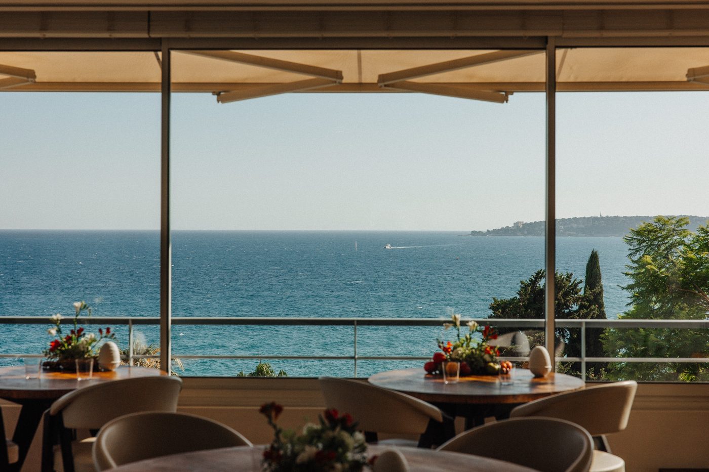 La salle du Mirazur, tables rondes fleuries devant une baie vitrée ouverte sur la Méditerranée et un cap boisé