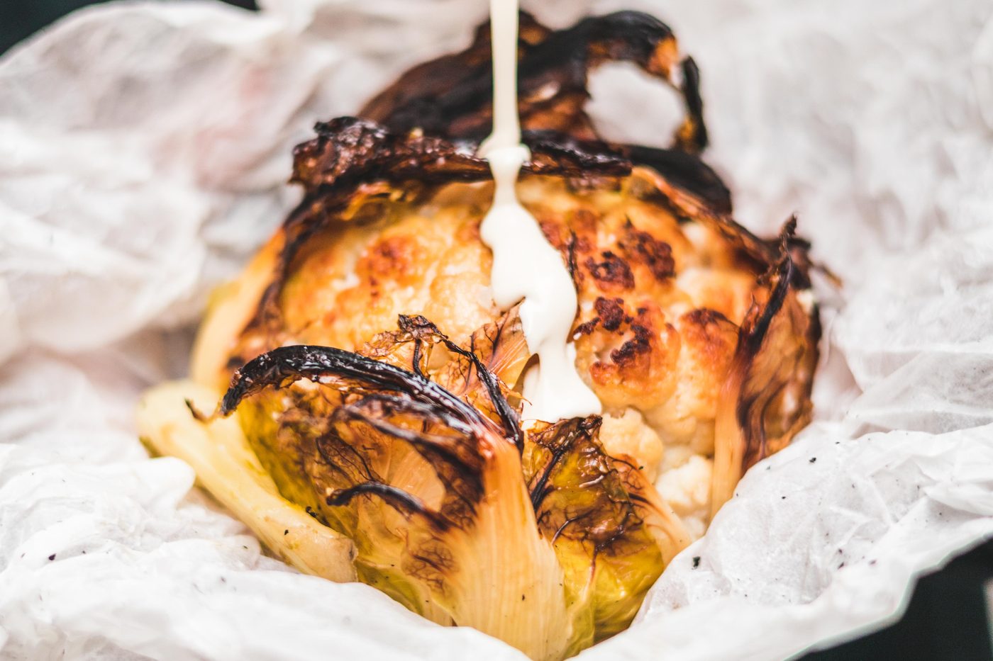 Un chou-fleur entier rôti, bruni et noirci, dans un papier blanc froissé, arrosé d'une sauce blanche