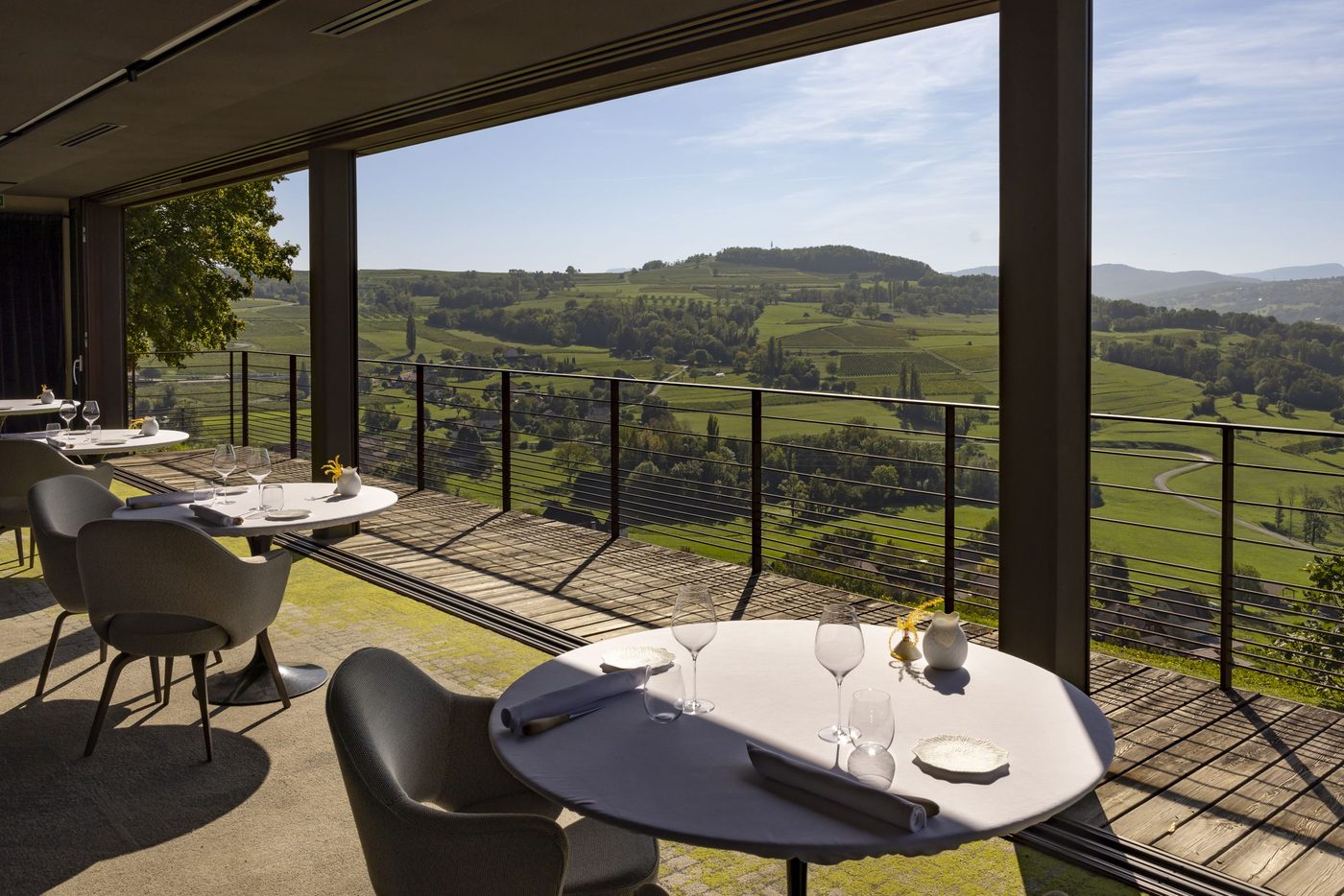 La salle des Morainières : tables rondes, fauteuils gris et grandes baies ouvertes sur une terrasse en bois et les coteaux