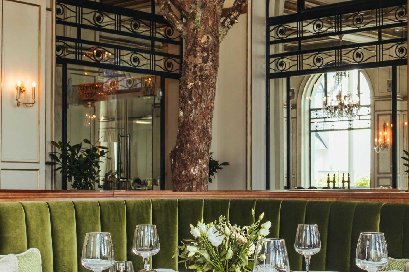 La salle du Pavillon, banquette de velours vert, verres sur les tables, un arbre et des lustres derrière les verrières