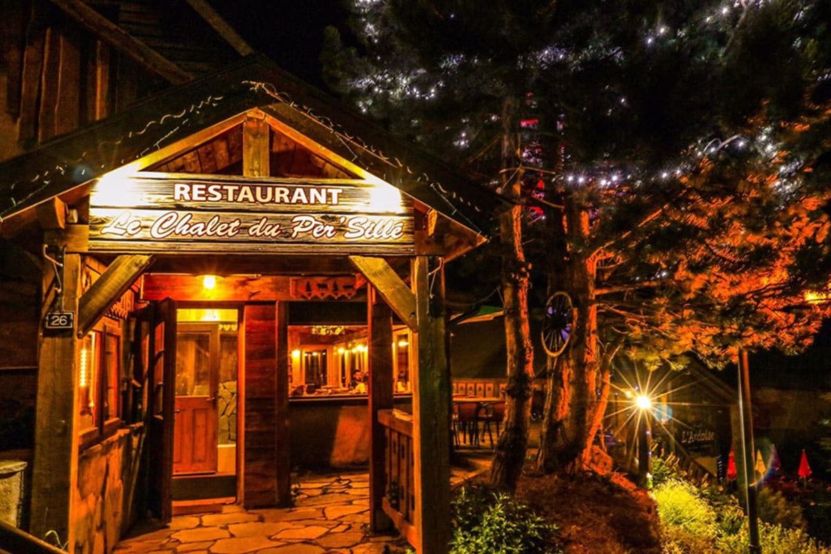 L'entrée en bois du Chalet du Pèr'Sillé la nuit, enseigne éclairée et guirlandes lumineuses dans les arbres