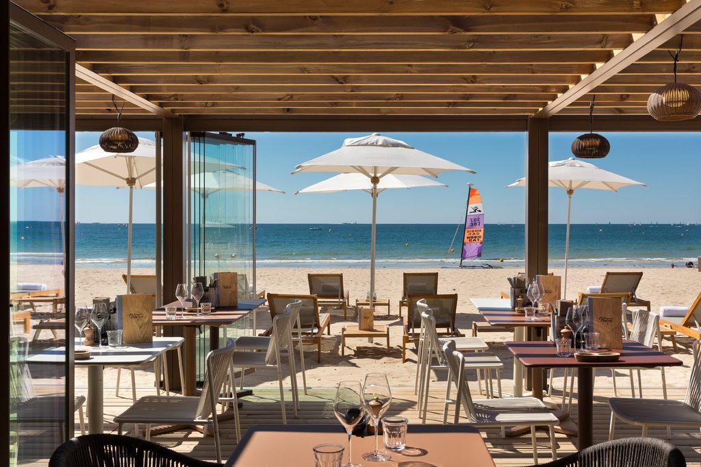 La terrasse du Ponton sous une pergola de bois, tables face à la plage et aux parasols blancs