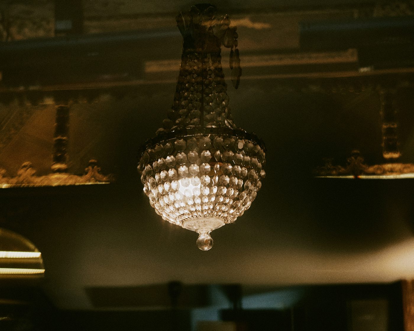 Un lustre à pampilles de cristal allumé sous un plafond doré, chez Racines
