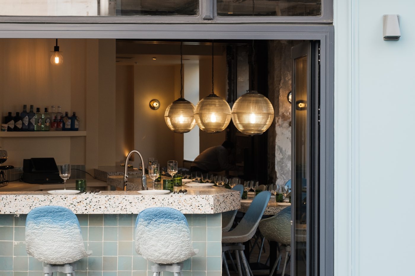 Le comptoir carrelé de Sables, tabourets de céramique bleue et suspensions en verre, vu par la devanture ouverte