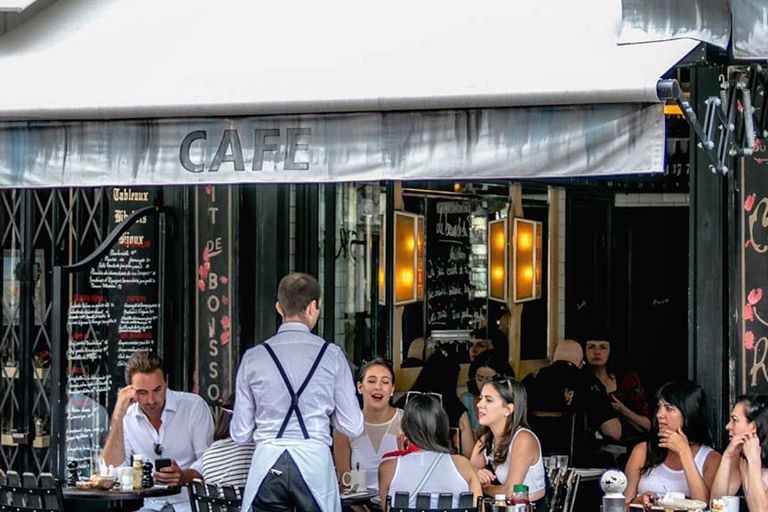 La terrasse du Saint-Régis sous son store blanc, un serveur en tablier et des clients attablés