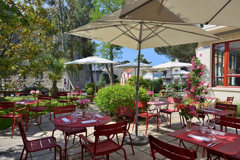 La terrasse fleurie de La Table du Saint-Christophe, tables et chaises rouges sous les parasols, au milieu des arbres