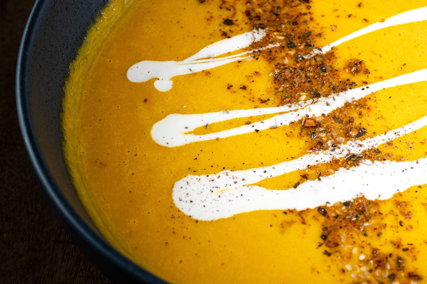 Un velouté orangé de Soya, servi dans un bol noir, avec un filet de crème végétale et des graines