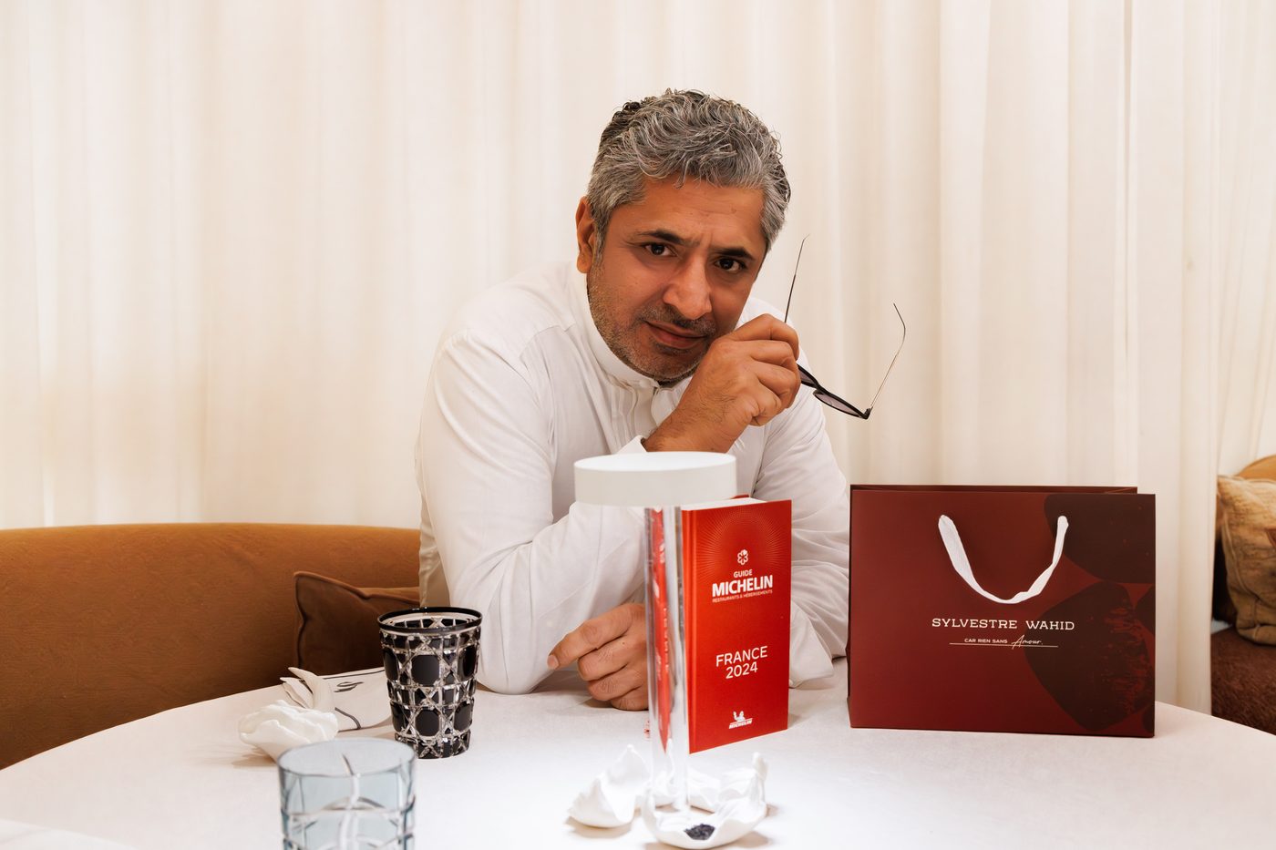 Sylvestre Wahid en veste blanche, attablé derrière un guide Michelin rouge et un sac à son nom