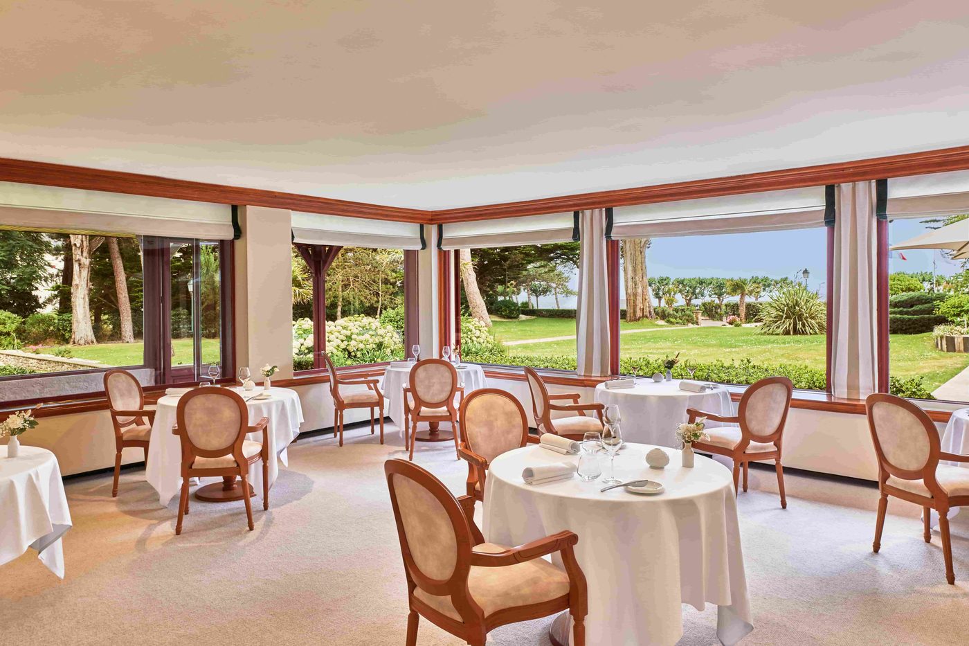 La salle de La Table du Castel, tables rondes nappées de blanc et fauteuils médaillon, grandes baies sur le parc