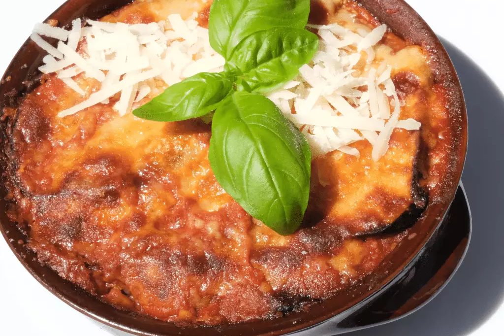 Une parmigiana d'aubergine gratinée dans un plat en terre, parsemée de parmesan et d'une feuille de basilic