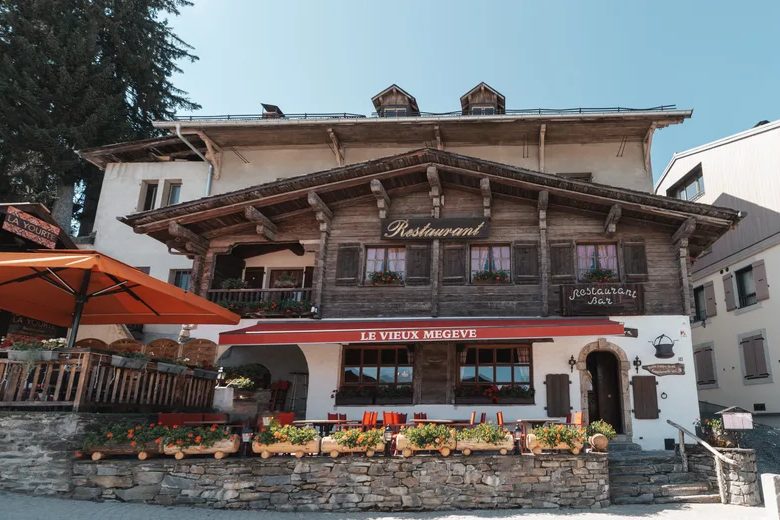 La façade du Vieux Megève, grand chalet de bois au store rouge, terrasse fleurie et parasols