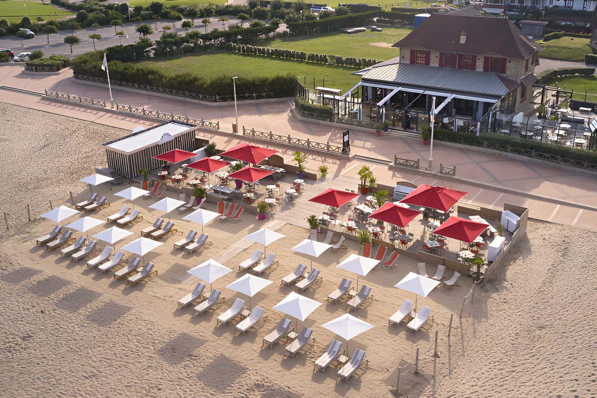 Vue aérienne de La Villa sur la Plage, la maison, ses parasols rouges et blancs et ses transats sur le sable