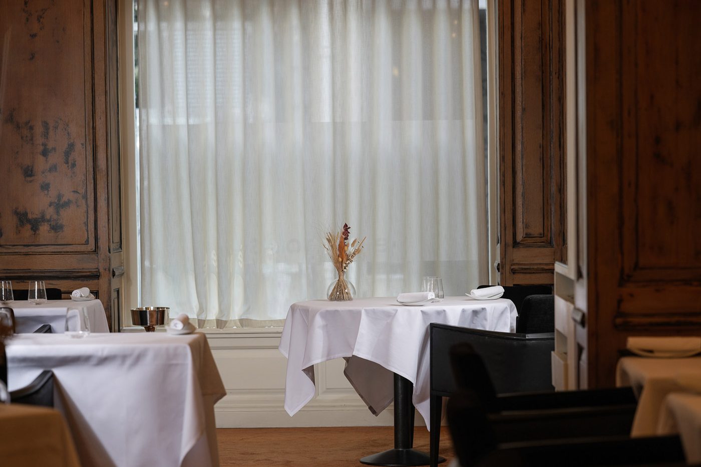 Une table nappée de blanc devant une grande fenêtre voilée, entre des boiseries anciennes, chez Zostera